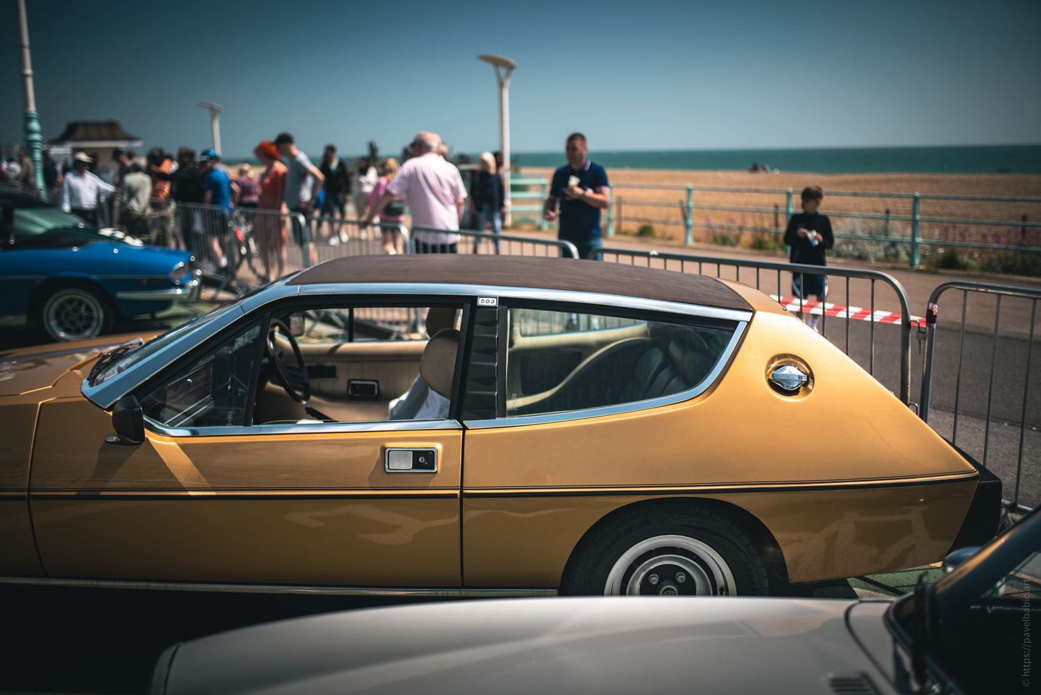 A vintage yellow car parked by the beach, with people milling around in the background on a sunny day.