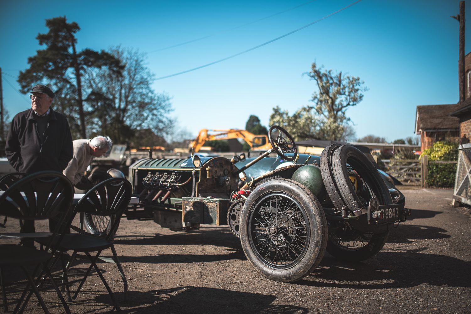 A vintage car with exposed mechanics sits in an outdoor setting, while two people engage nearby under a clear blue sky.