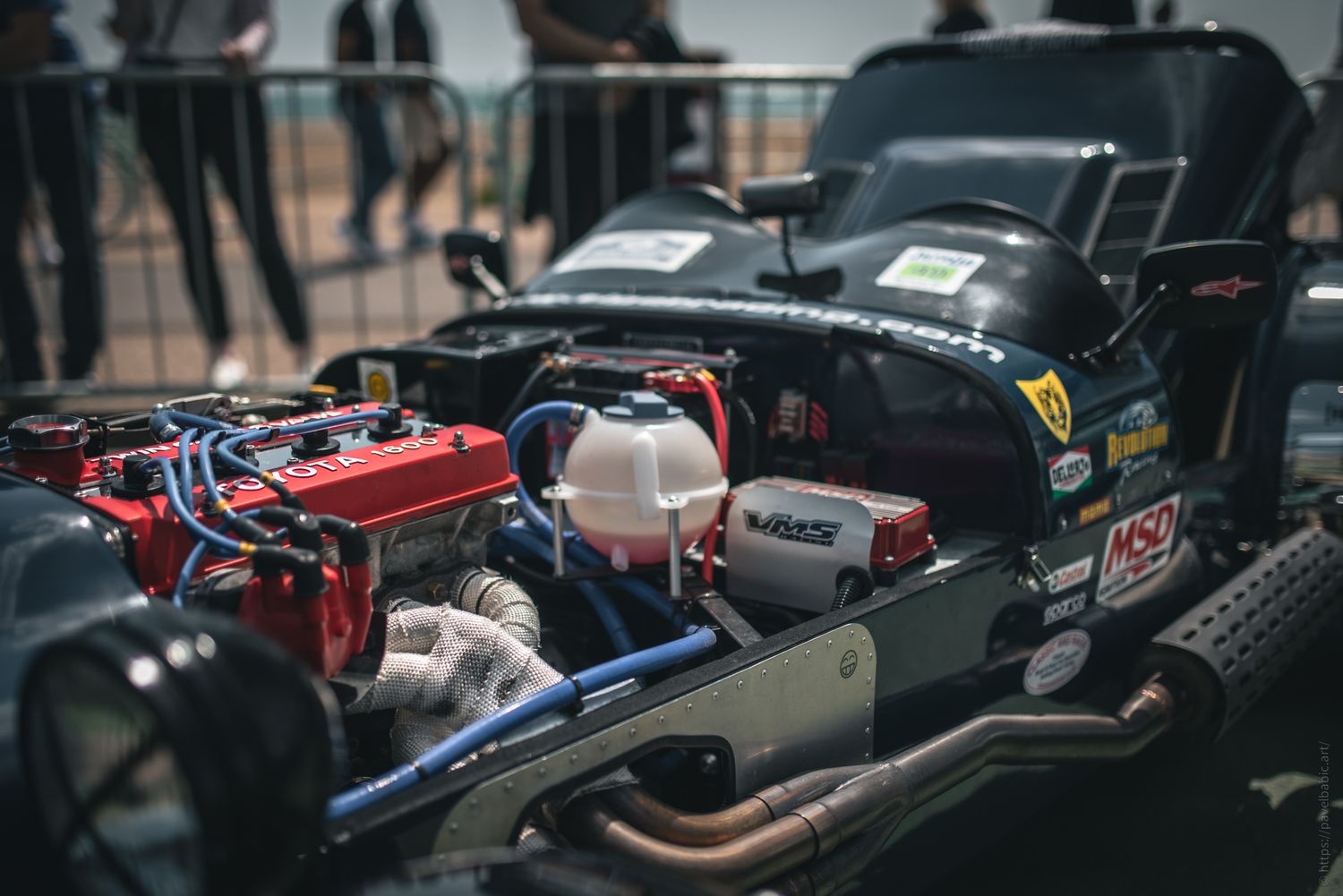 Close-up of a racing car's engine compartment, showcasing colorful wiring, a red battery, and intricate mechanical components.