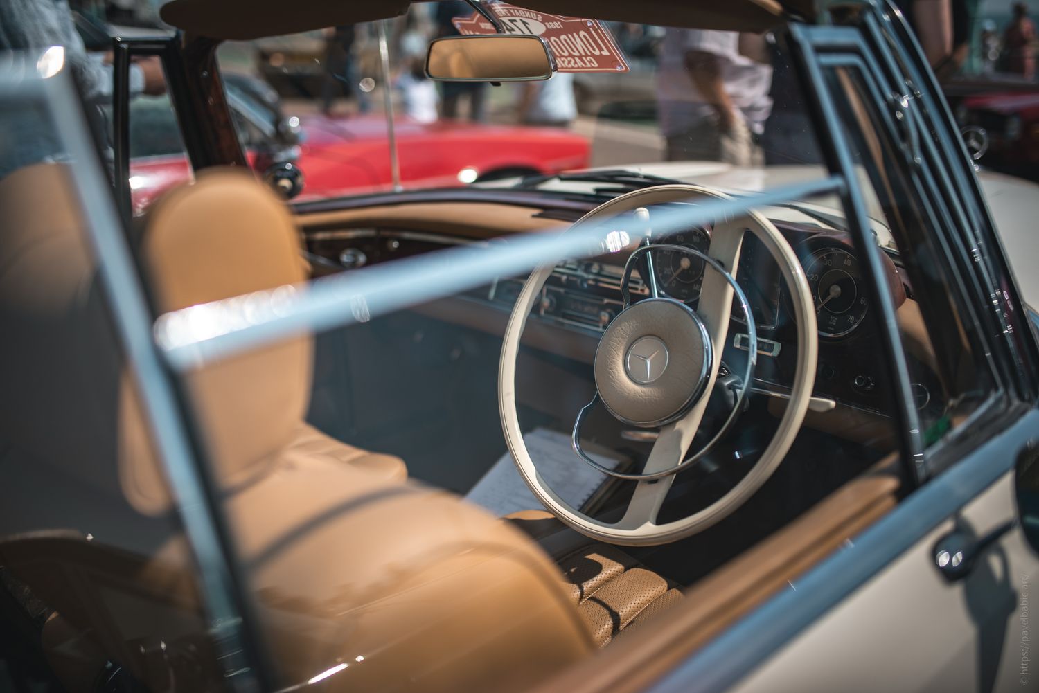 Vintage car interior featuring a tan leather seat, classic steering wheel, and dashboard, with an outdoor event ambiance.