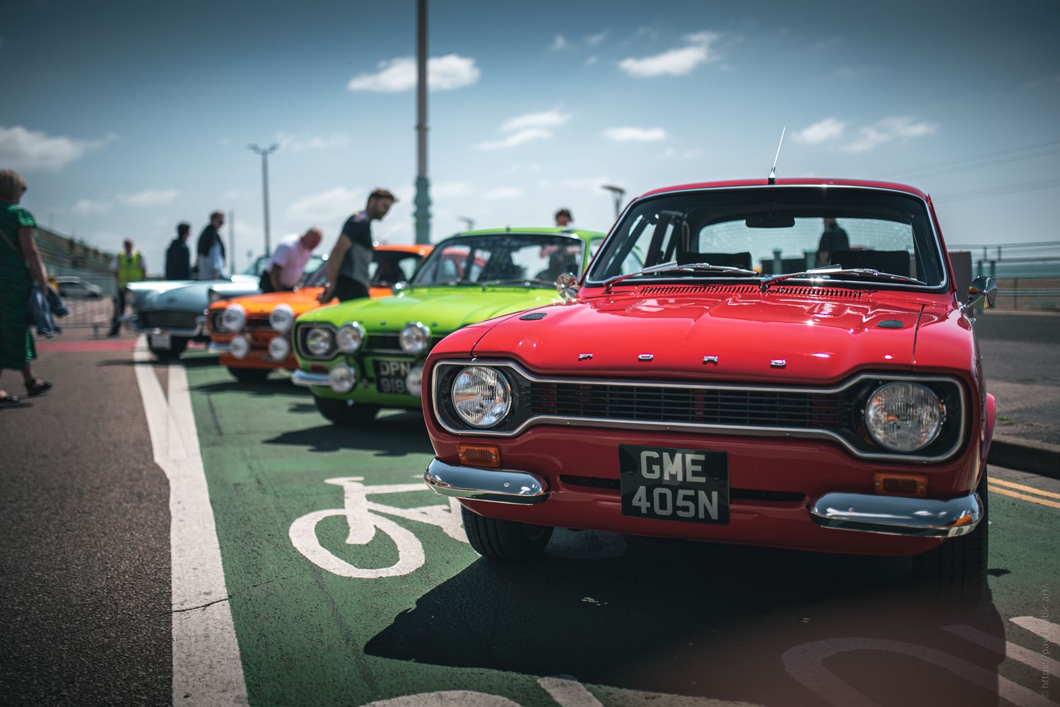 A vivid red vintage car is parked in front, with colorful classic cars lined up behind it, against a sunny seaside backdrop.