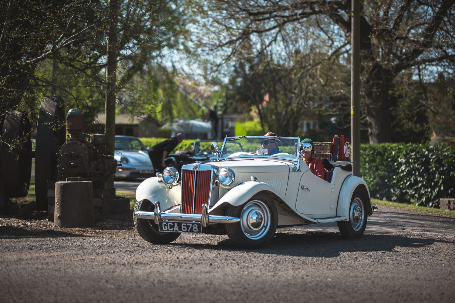 A vintage white convertible car parked on a gravel road, surrounded by trees and greenery, with two people enjoying the ride.