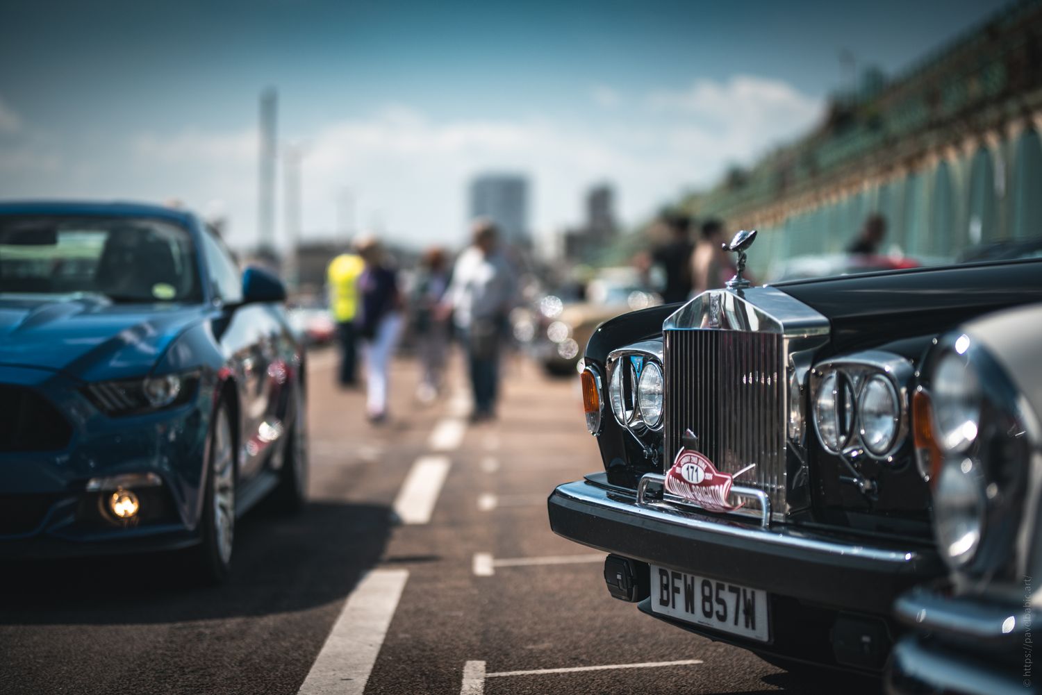 Close-up of a vintage Rolls-Royce and a blue sports car at a car event, with people and city buildings in the background.