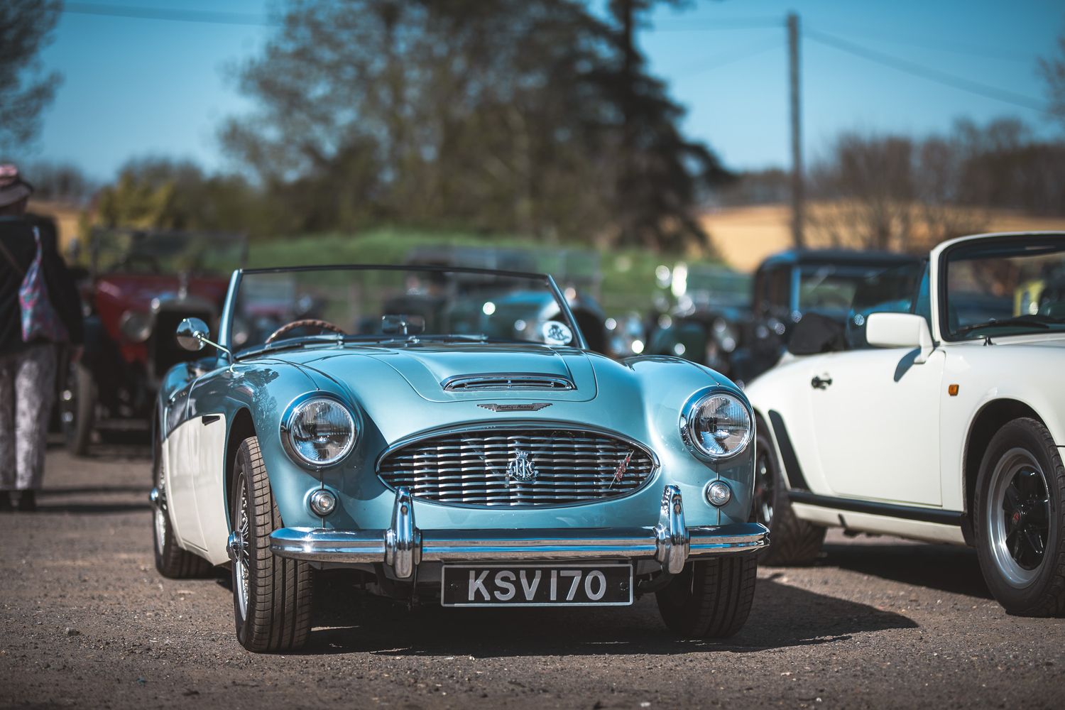 A classic blue sports car parked at a vintage car event, with a license plate KSV170, surrounded by other vehicles in the background.