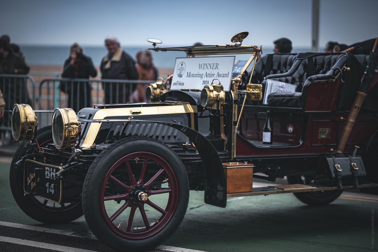 A vintage car with a brass and black finish, showcasing its elegant design, parked at a seaside event with spectators in the background.