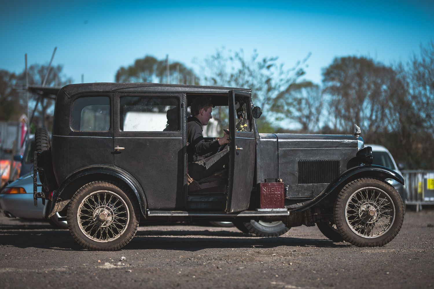 A vintage 1920s black car with a driver at the open door, parked in a sunny outdoor setting with trees in the background.