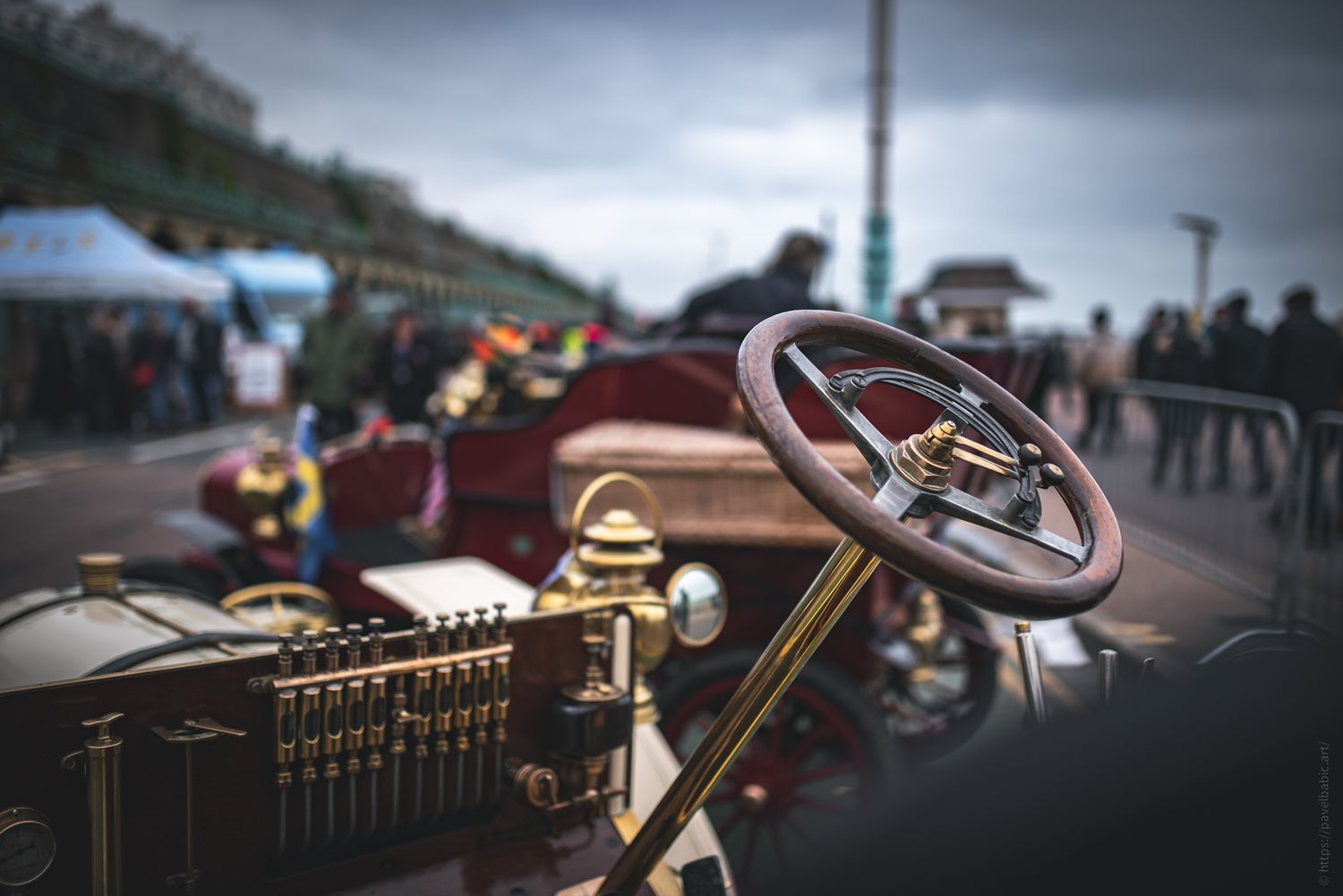 Close-up of a vintage car's wooden steering wheel with gleaming brass details, set against a bustling outdoor event scene.