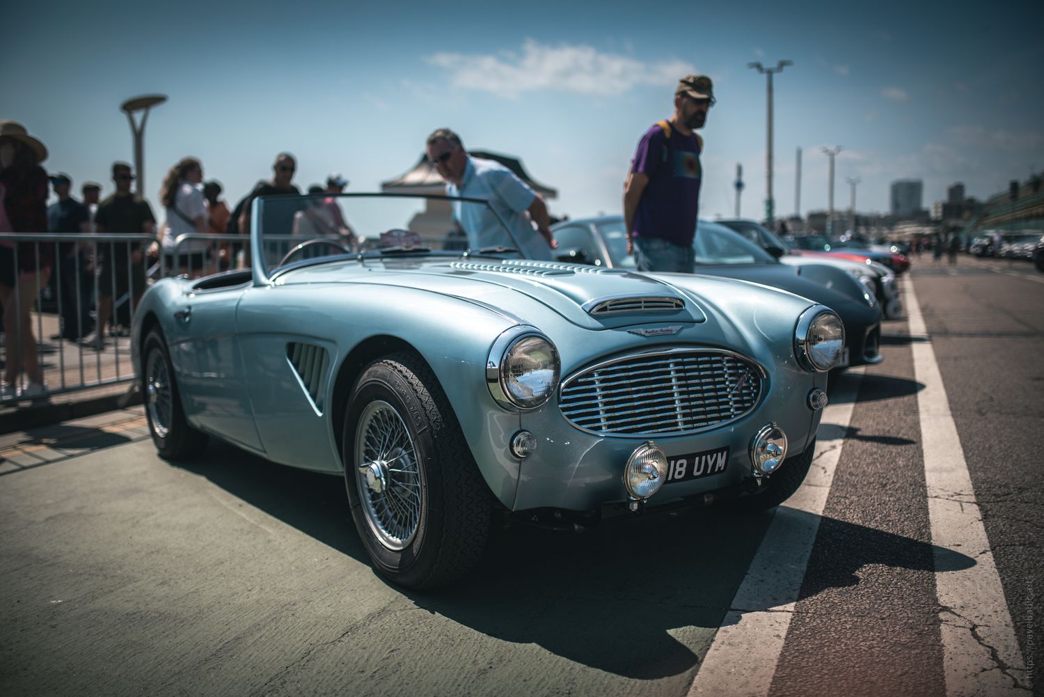 A classic blue convertible sports car parked along a bustling promenade, with people admiring it under a sunny sky.