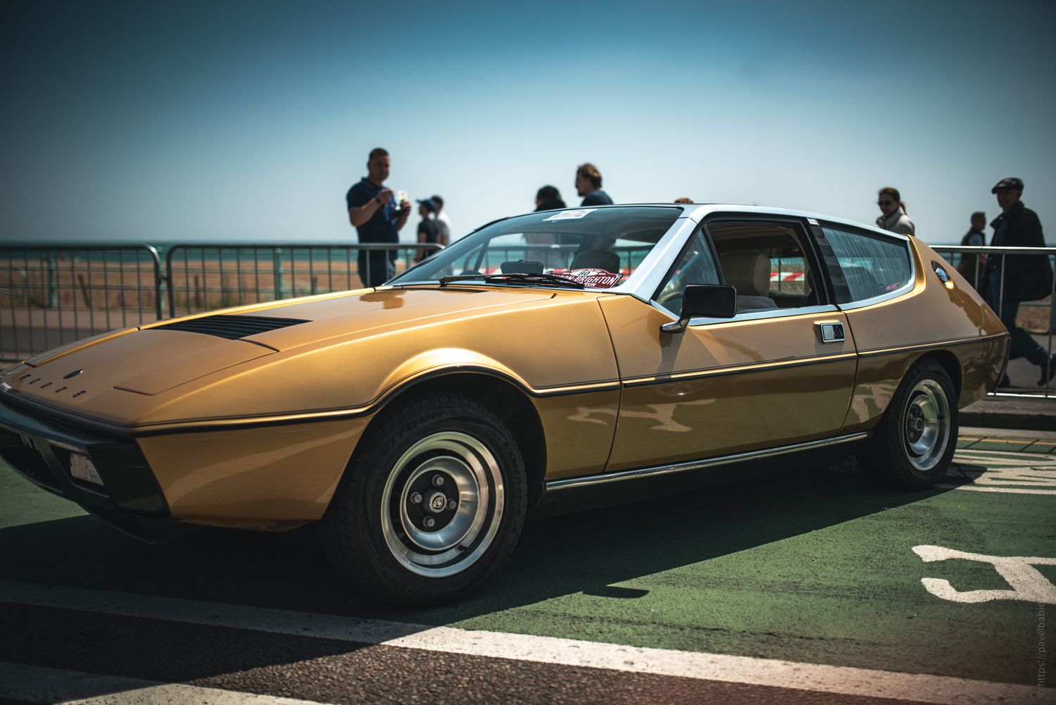A vintage gold sports car parked, with people admiring it in the background on a sunny day by the beach.