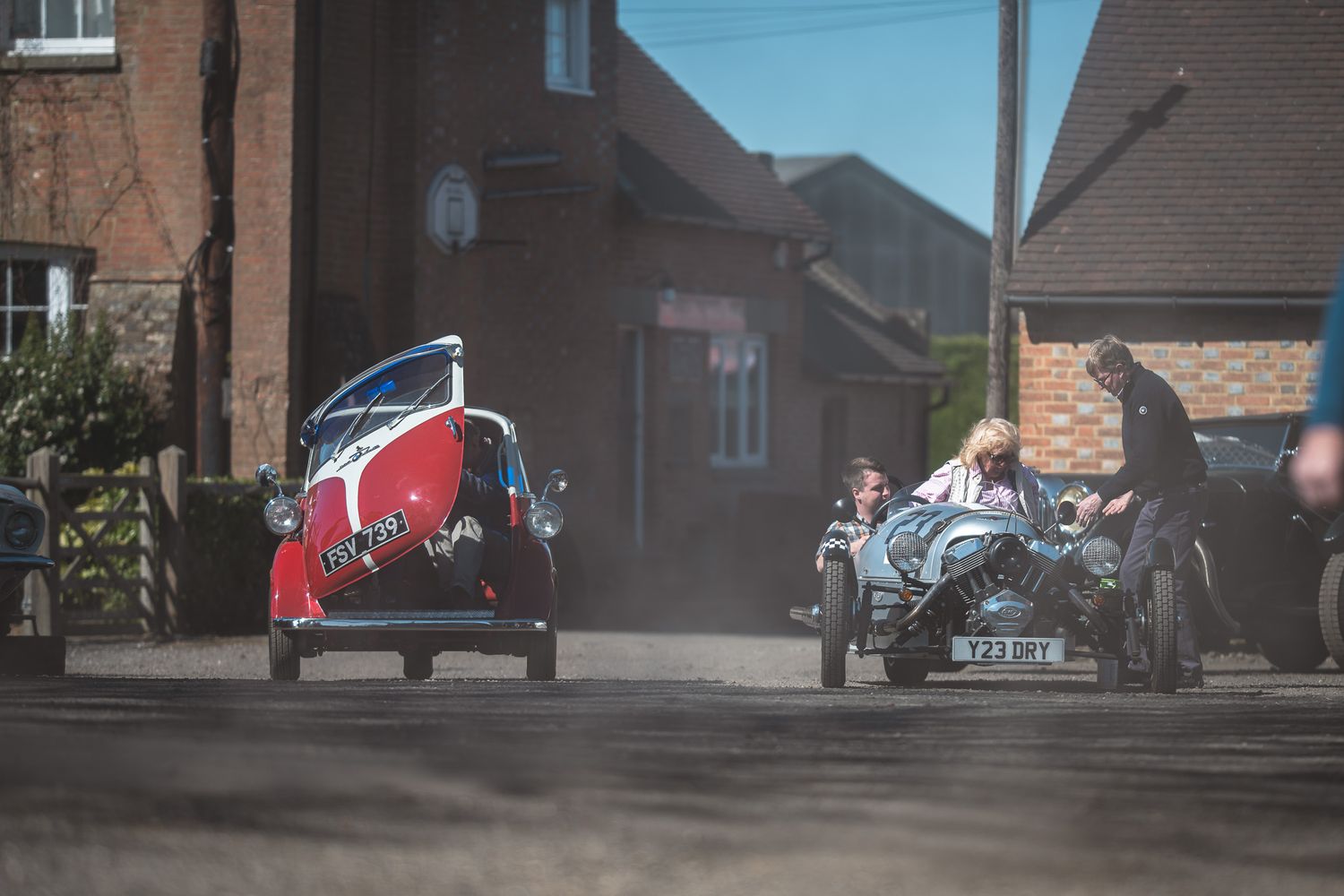 Two vintage cars, a red one with a unique design and a blue sports car, drive down a dusty road in a rustic village setting.