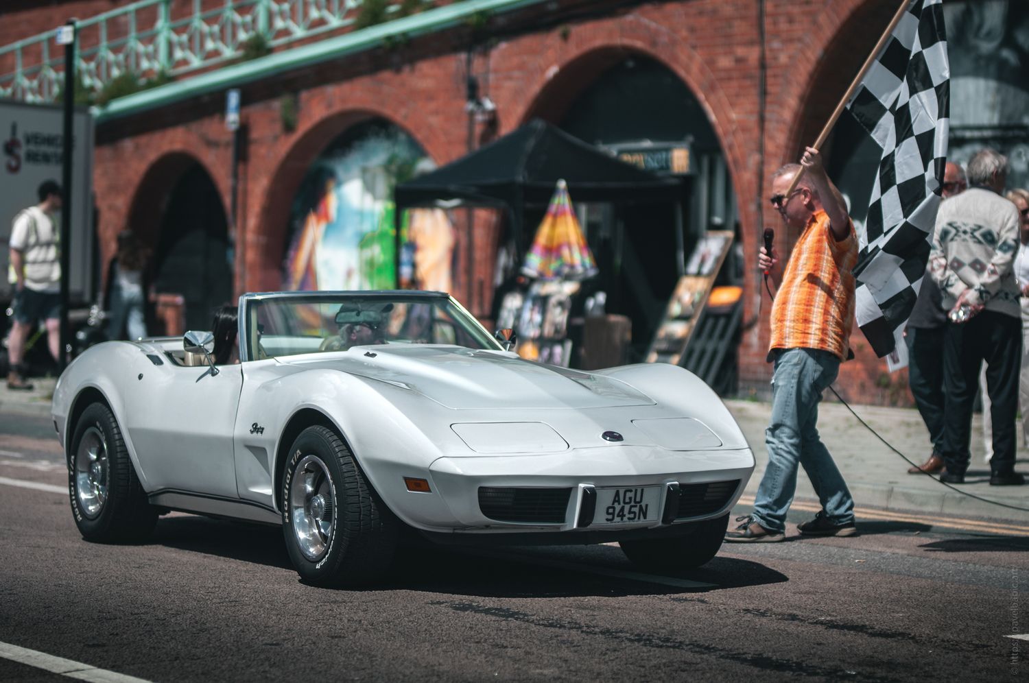 A silver convertible sports car drives past a man holding a checkered flag in a vibrant urban setting with brick buildings.