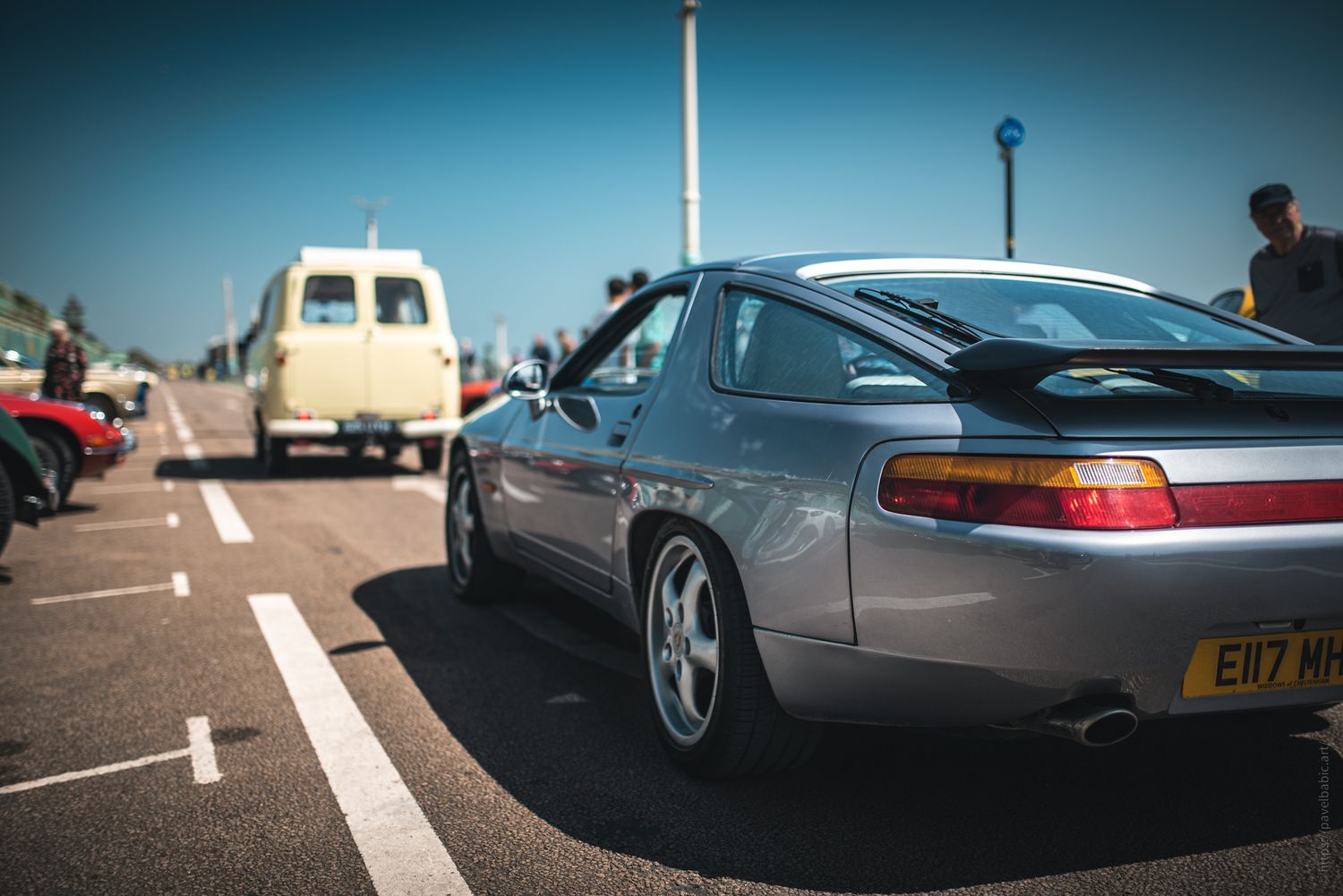 A silver Porsche 928 parked beside a vintage van at a car show, with attendees admiring nearby vehicles under a clear blue sky.