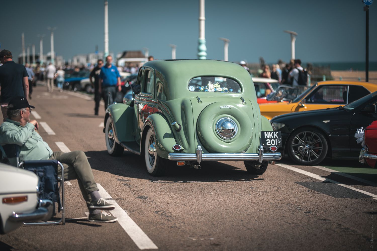 A vintage green car parked on a lively promenade, surrounded by spectators and classic cars under a clear blue sky.