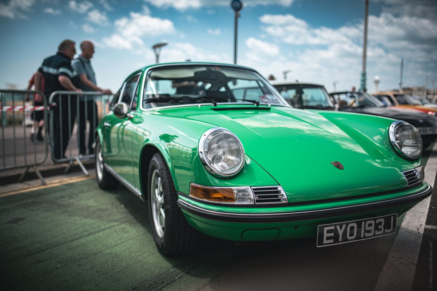 A vibrant green classic Porsche parked with onlookers in the background, showcasing its sleek design under a blue sky.