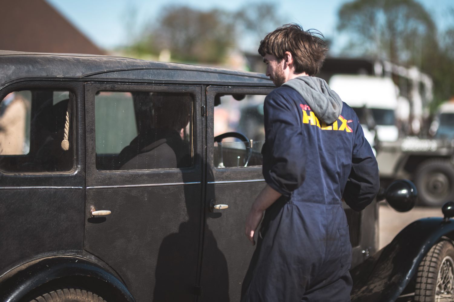 A young man in a blue jumpsuit examines an old black car, reflecting a vintage setting and a hands-on approach to restoration.