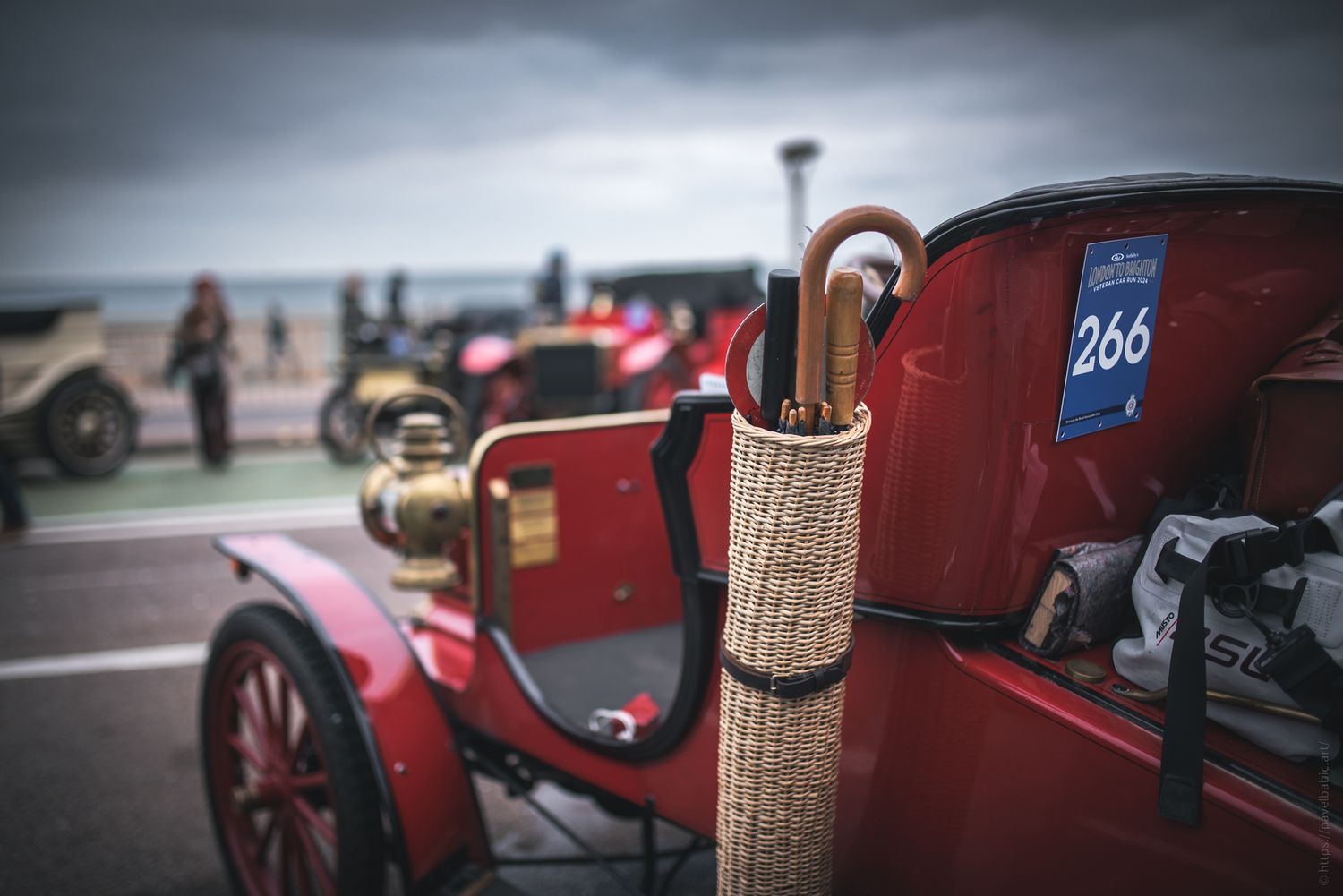 A vintage red car parked by the beach, featuring a numbered tag and an umbrella neatly stored in a side holder.