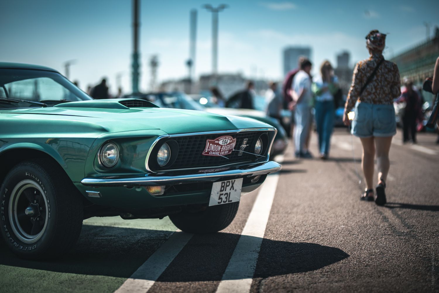 A classic green muscle car parked on a sunny street with people walking by, capturing a vibrant automotive gathering atmosphere.