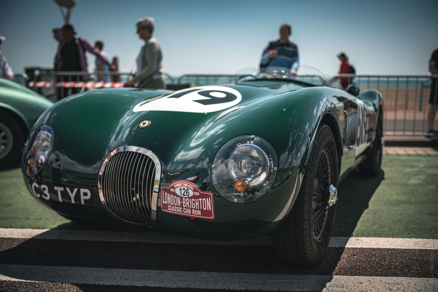 A classic green racing car with number 19, parked at an event, surrounded by spectators and vintage cars in the background.