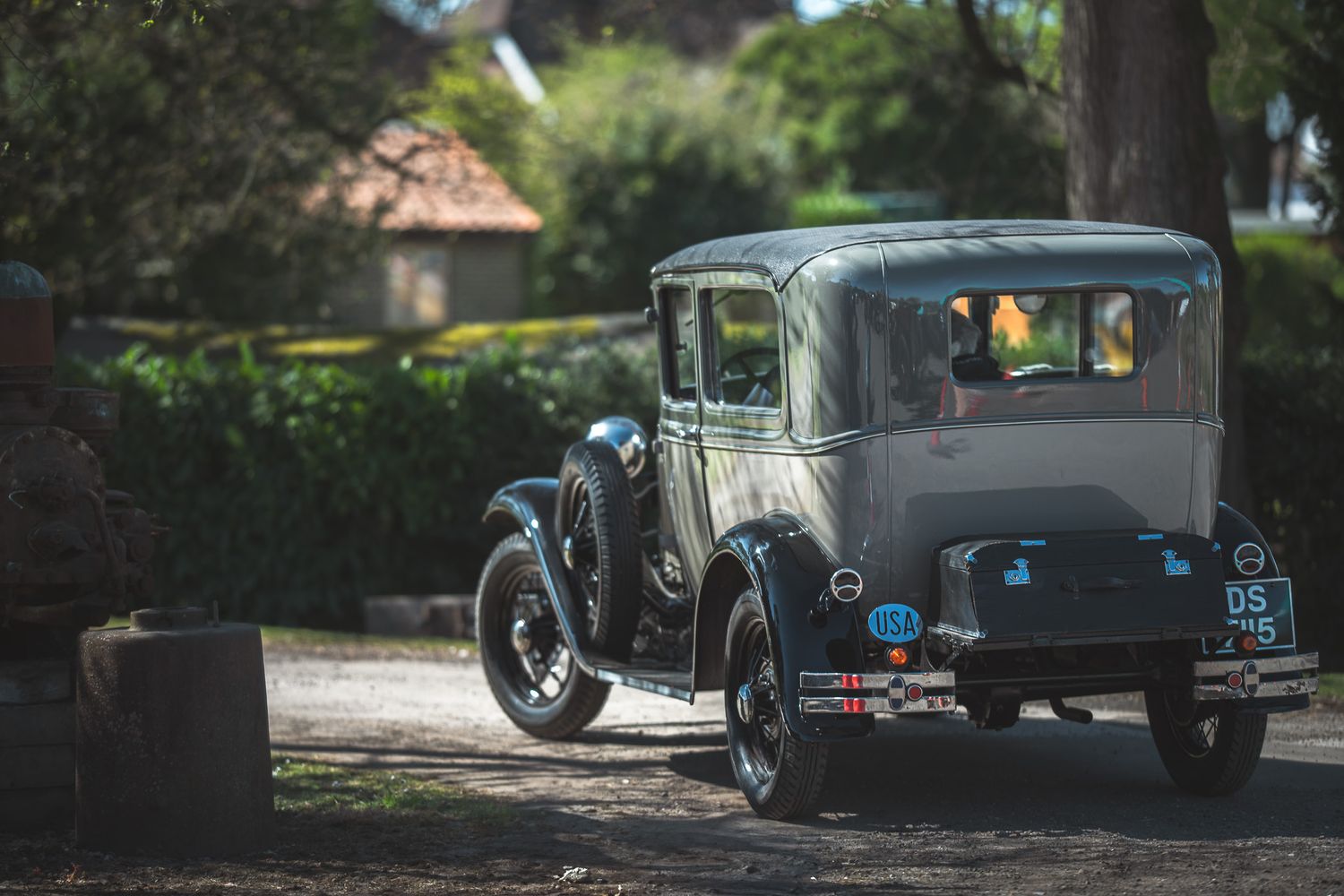 A vintage gray car with a rounded roof drives on a sunlit road surrounded by lush greenery. The scene evokes nostalgia and tranquility.