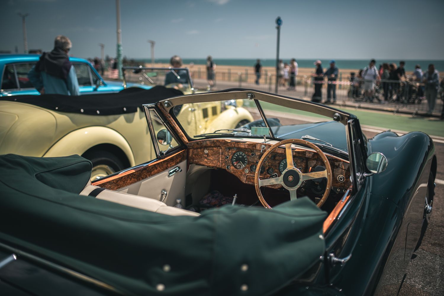 Interior view of a classic convertible car with a wooden dashboard, parked by the beach, with onlookers in the background.