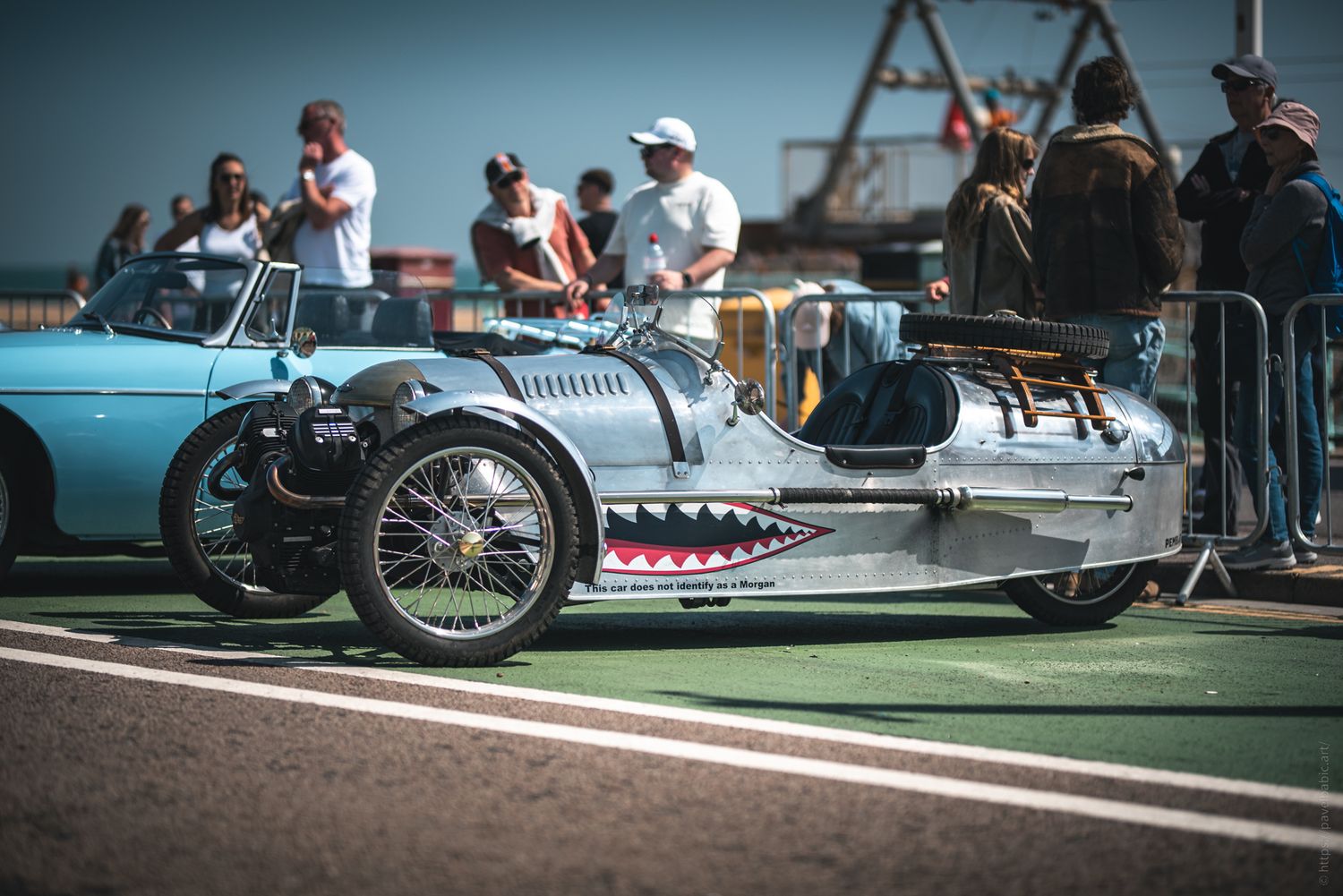A vintage silver three-wheeled car with a shark-mouth design sits at a lively outdoor event, surrounded by onlookers and attractions.