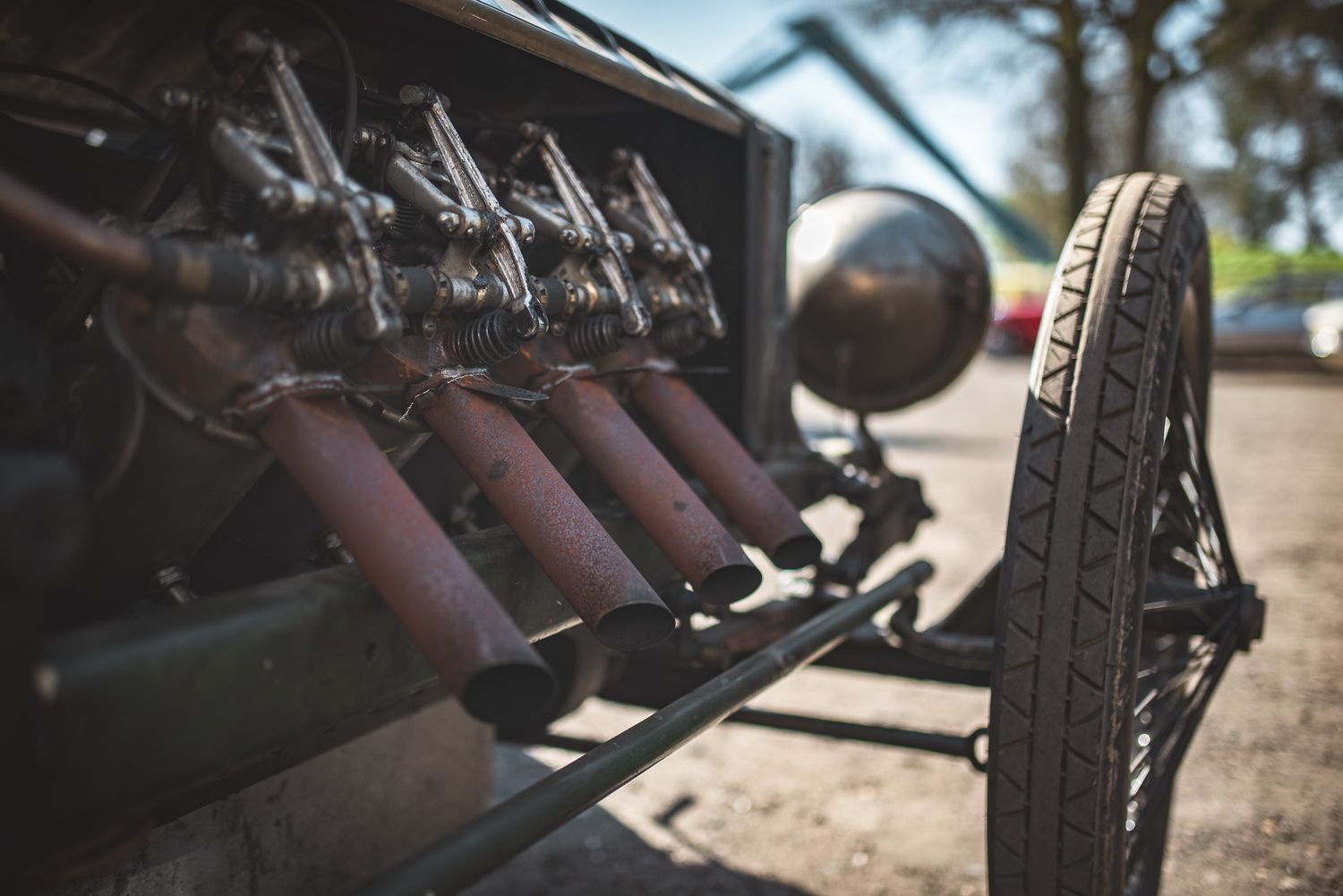 Close-up of an antique car's exposed engine with four exhaust pipes, showcasing vintage metalwork and a wooden wheel in the foreground.