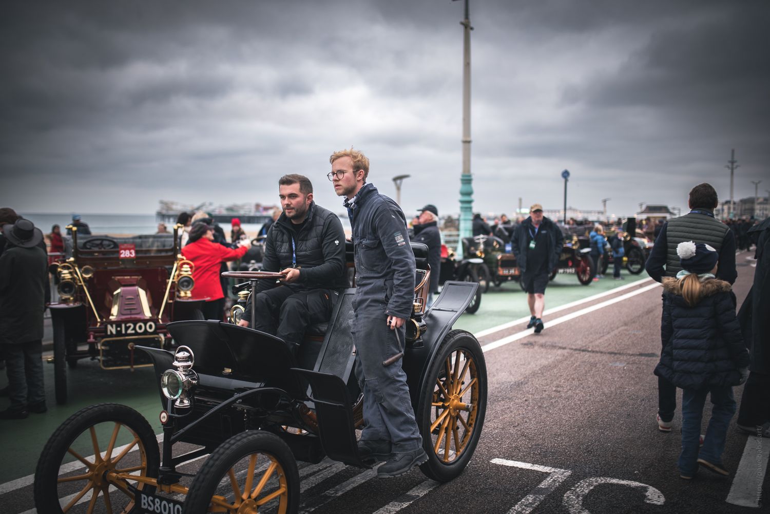 Two men in vintage attire drive a classic car along a promenade, surrounded by onlookers and antique vehicles under cloudy skies.