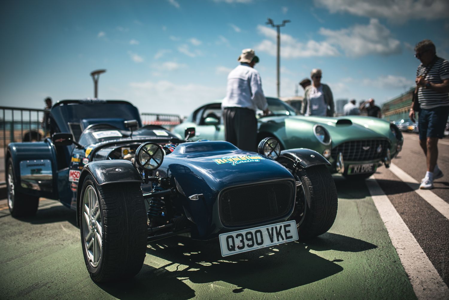 A blue vintage sports car parked along a seaside promenade, with people admiring it and another classic car nearby.