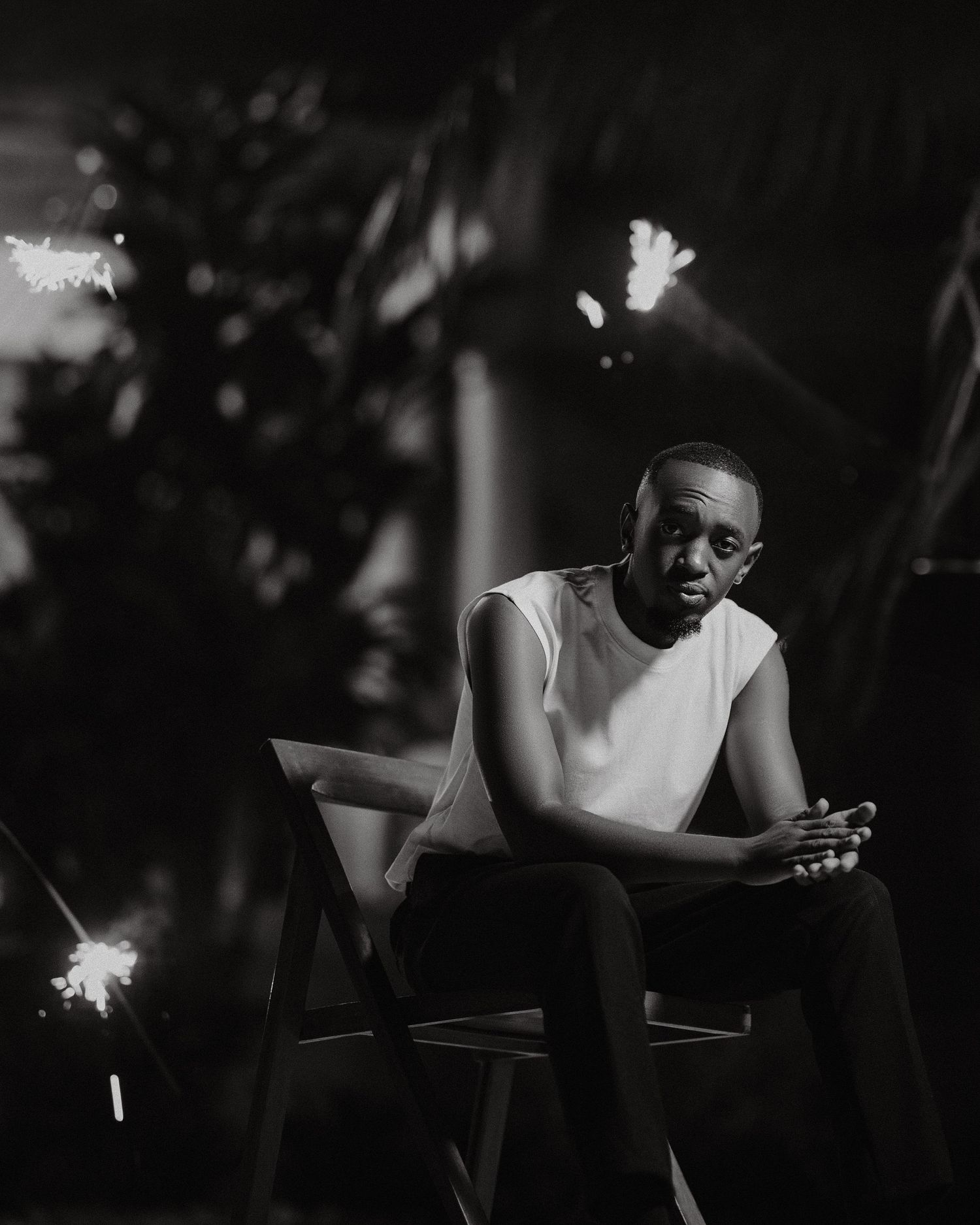 Black and white artistic portrait of a person sitting thoughtfully in a white sleeveless shirt against a dark background.