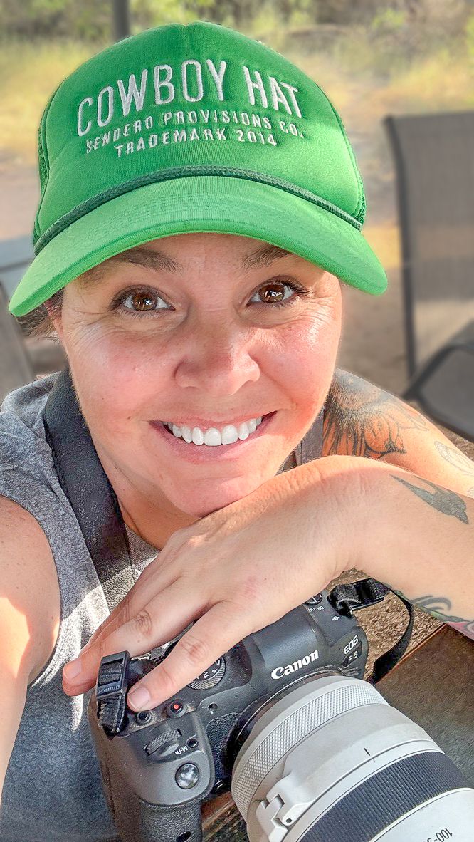 Photographer wearing a bright green Cowboy Hat branded baseball cap holds a Canon camera with telephoto lens.