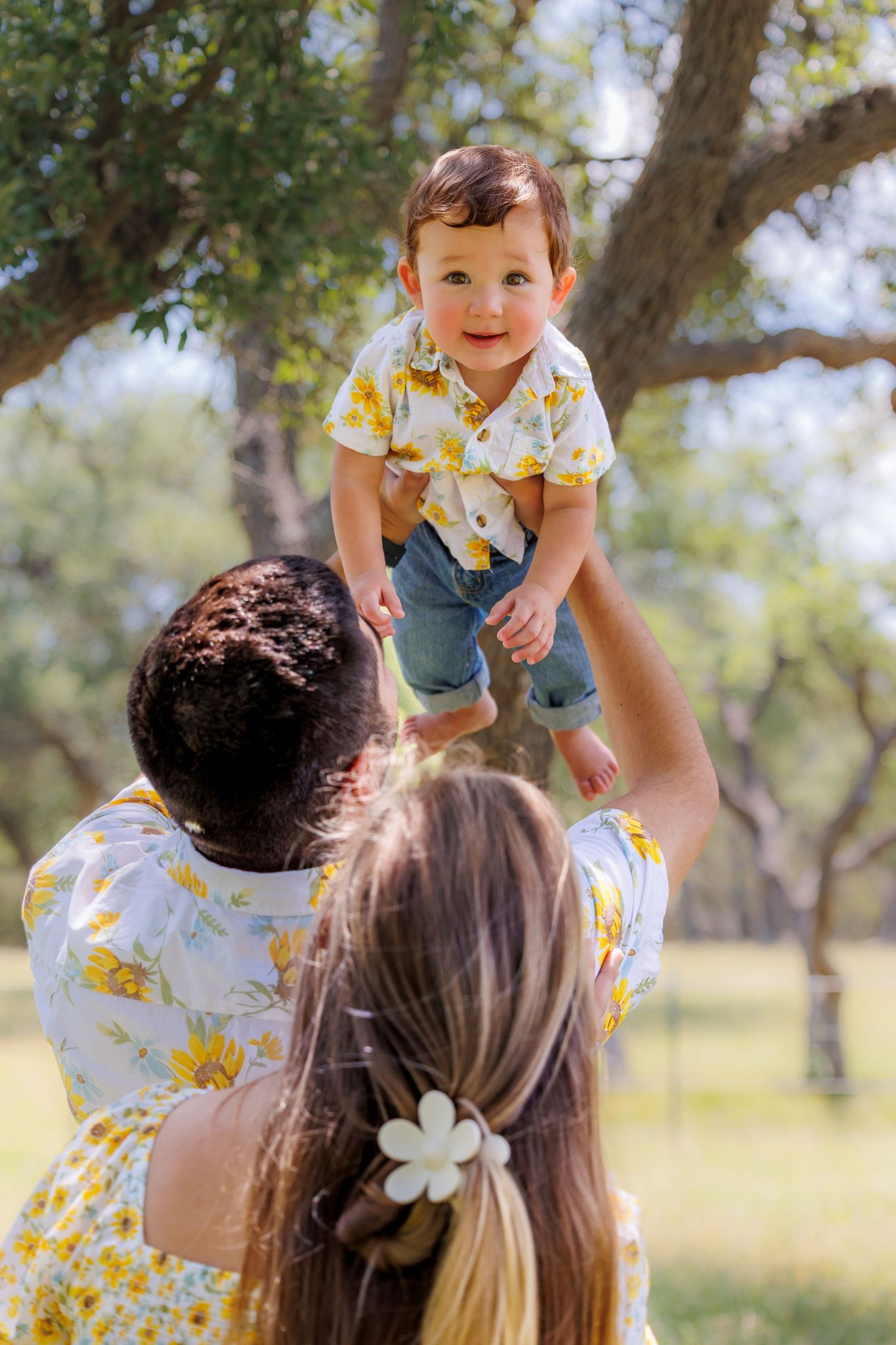 A joyful moment as a small child in yellow floral shirt and denim shorts is lifted up high in a sunny park setting.