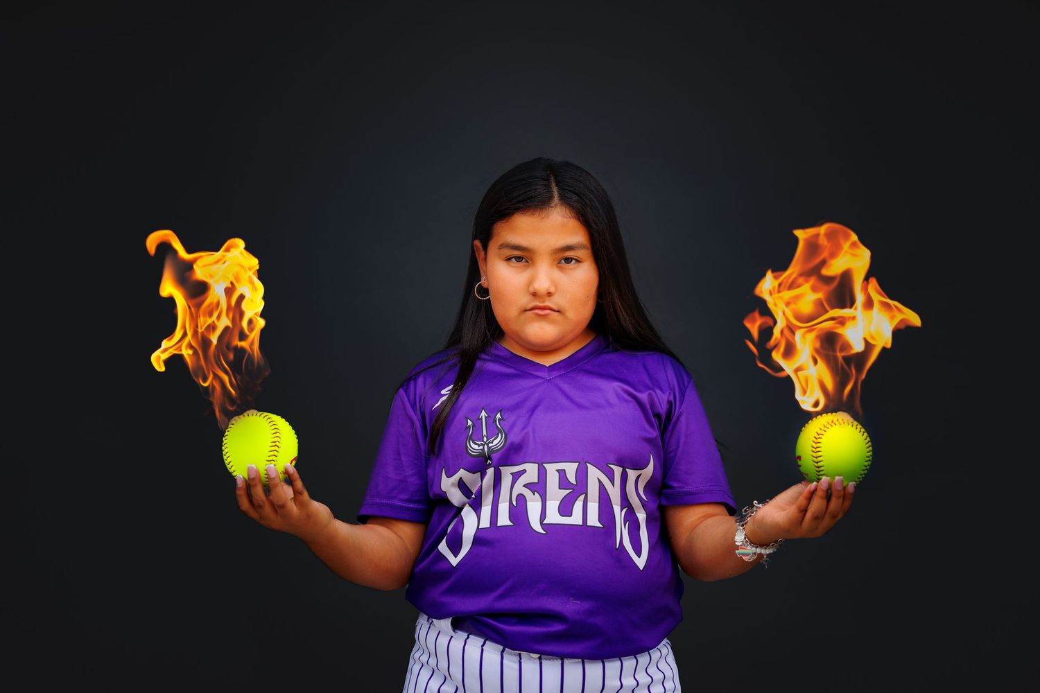 Baseball player wearing purple jersey holds up flaming tennis balls in dark setting for dramatic sports photography.