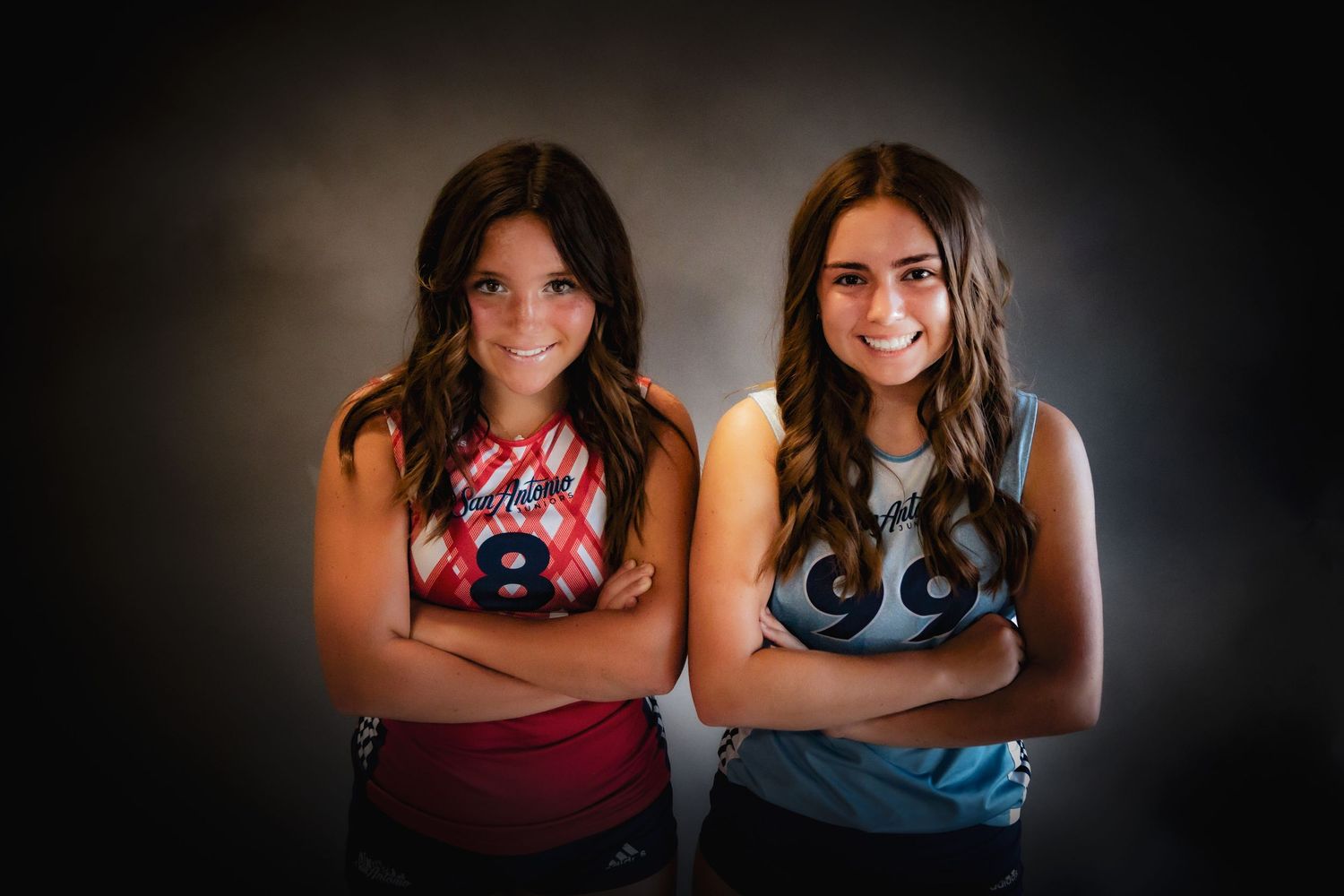 Two young athletes pose with arms crossed against dark background in sports uniforms.