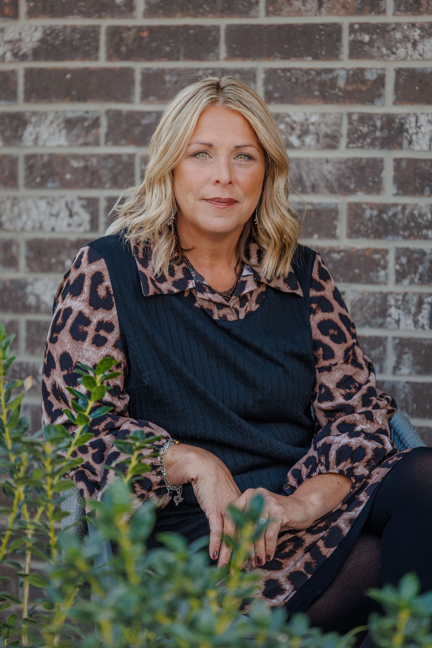 Woman in black dress with leopard print sleeves poses outdoors against brick wall with greenery.