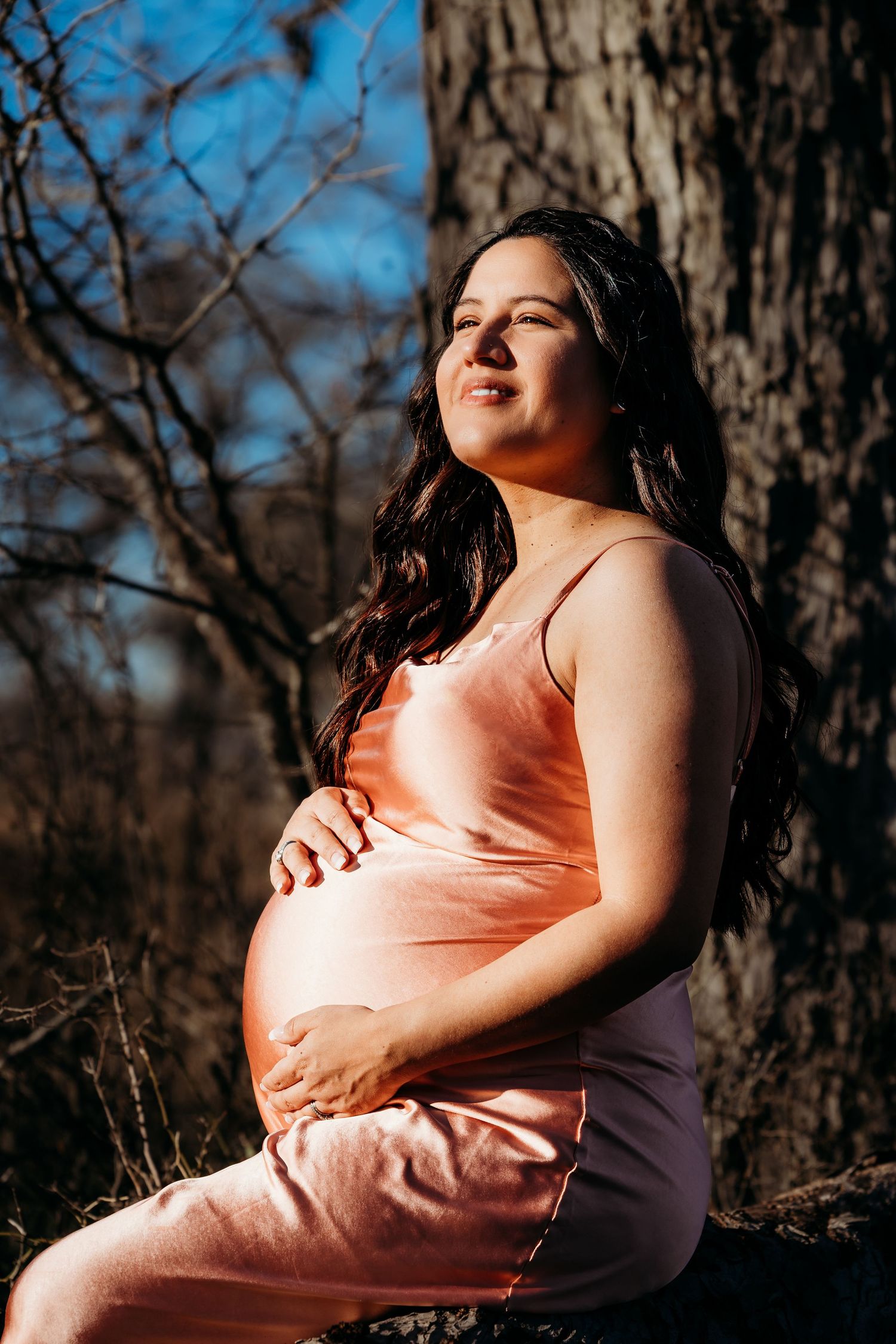 A pregnant woman in a peach-colored dress sits against a tree trunk during a sunny outdoor maternity photoshoot.