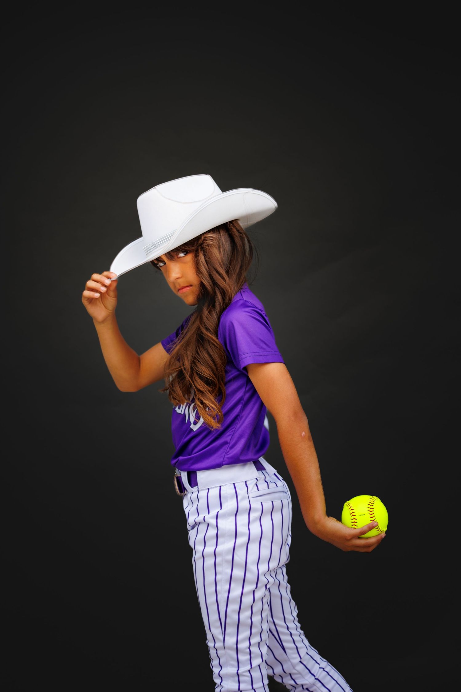 Softball player in purple jersey and pinstriped pants catches a yellow ball while wearing a white cowboy hat.
