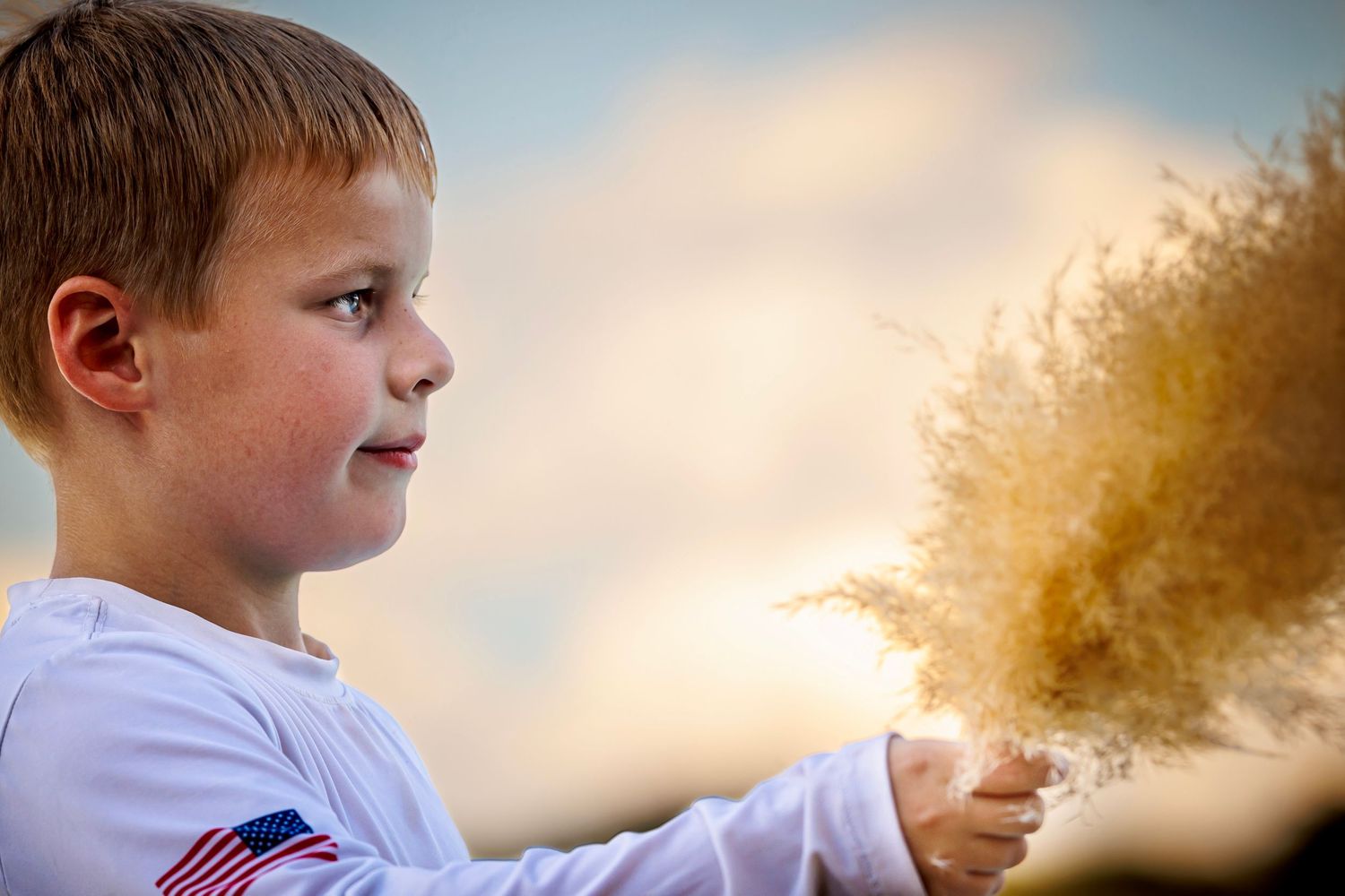 Child playing with floating dandelion seeds at sunset in a white shirt with American flag detail.