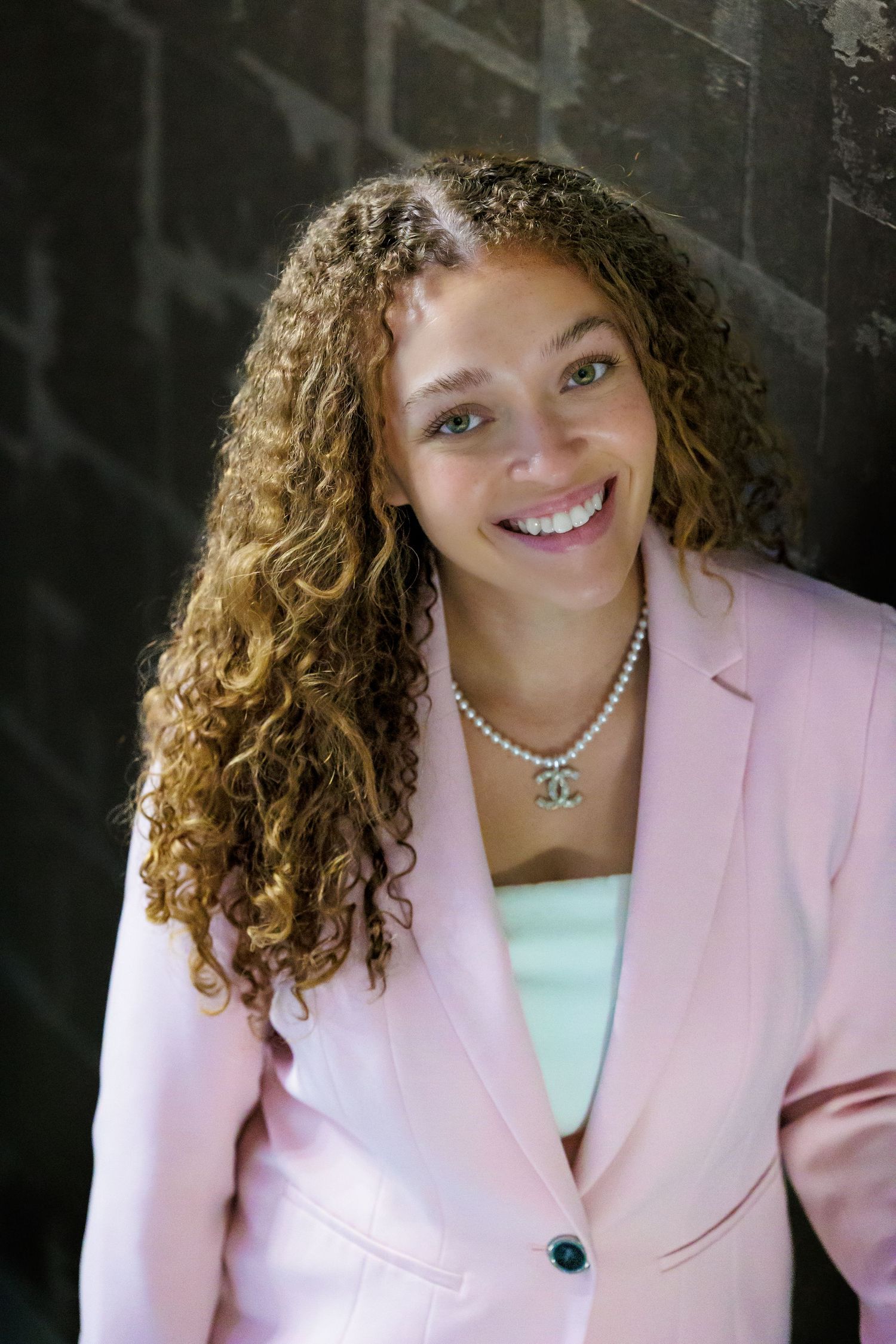 Professional headshot of someone with curly hair wearing a pink blazer and white top against a dark background.