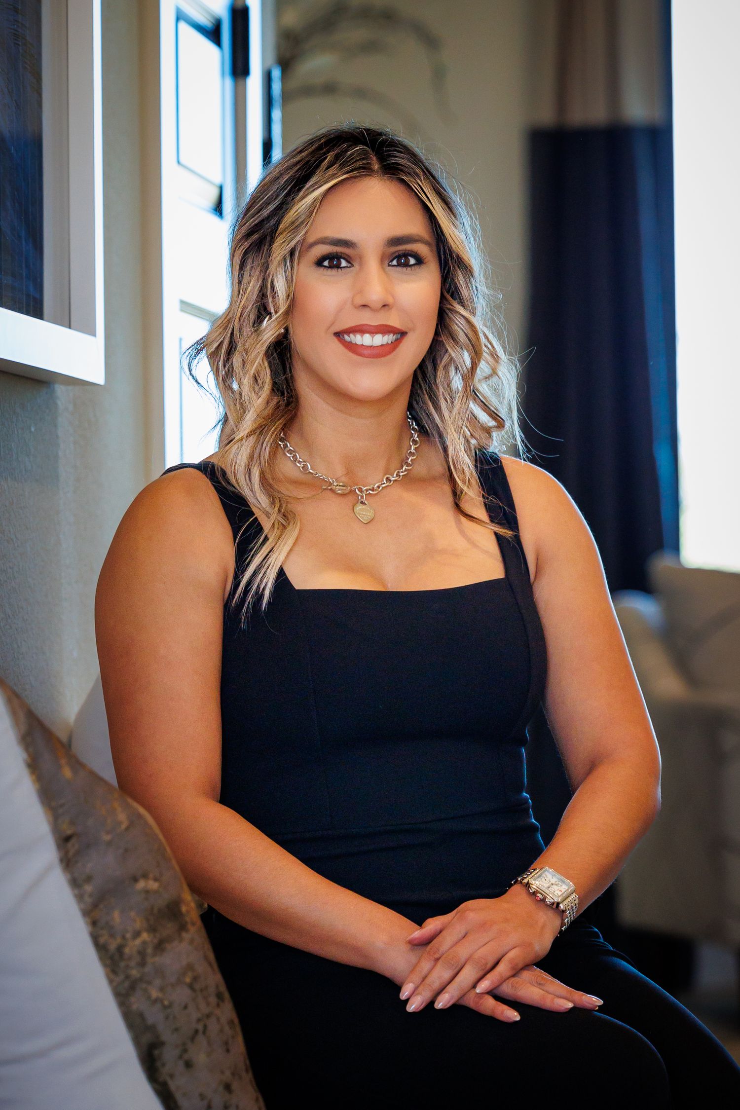 Woman in elegant black sleeveless dress poses indoors wearing silver necklace and wavy blonde hair.
