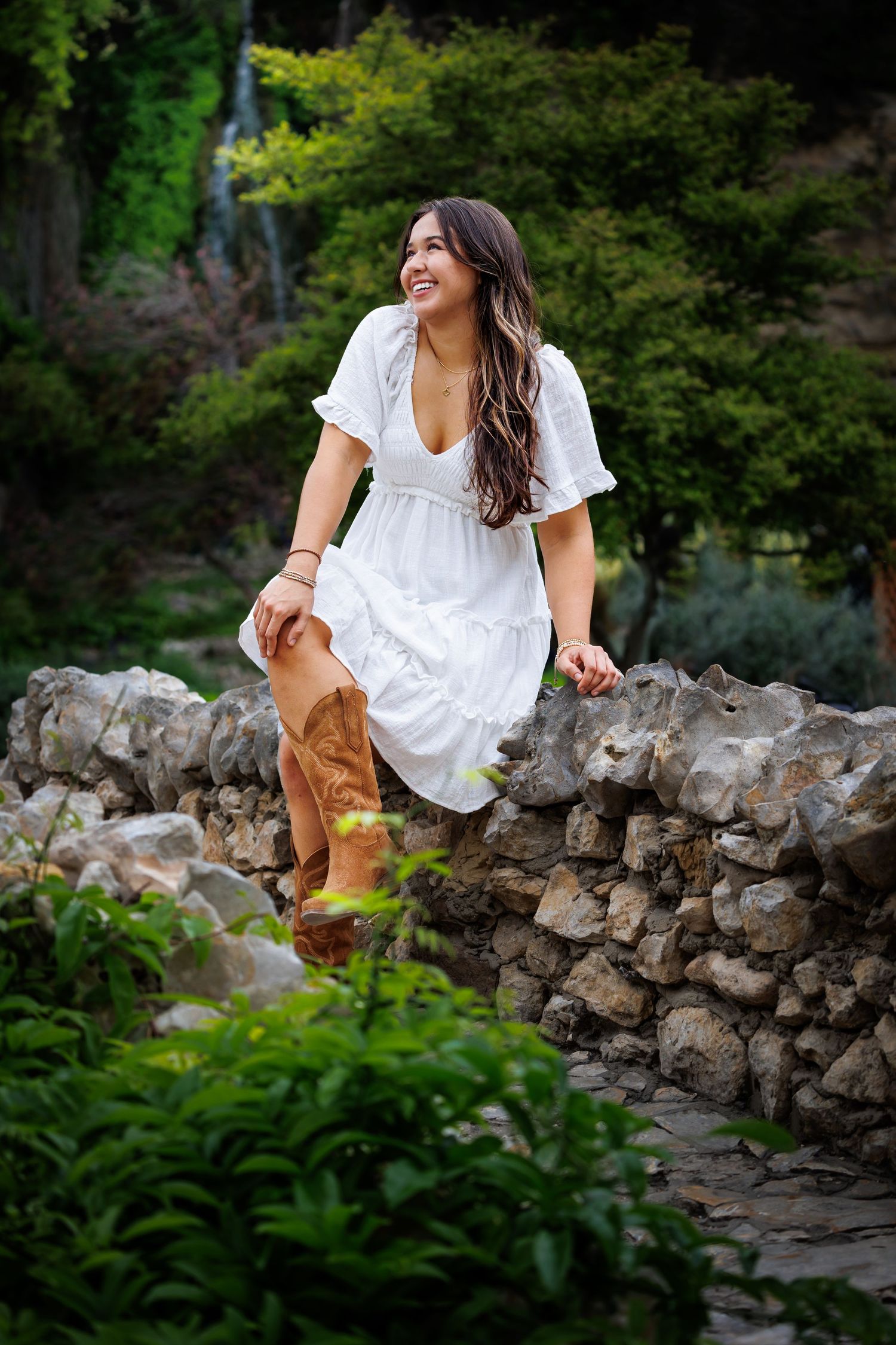 Woman in white summer dress and brown boots sitting on stone wall in garden setting.