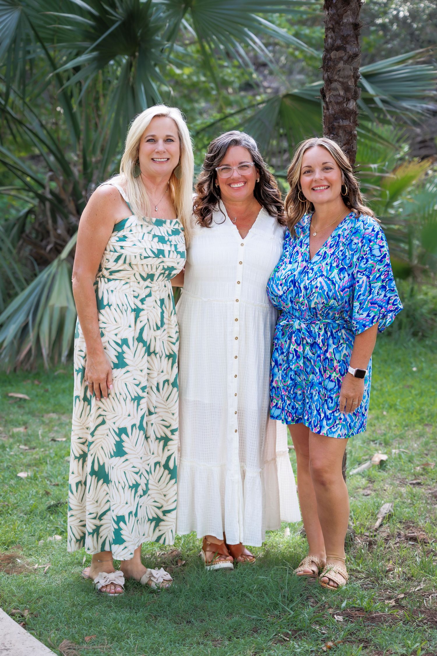 Three women in summer dresses pose together in front of tropical palm trees.