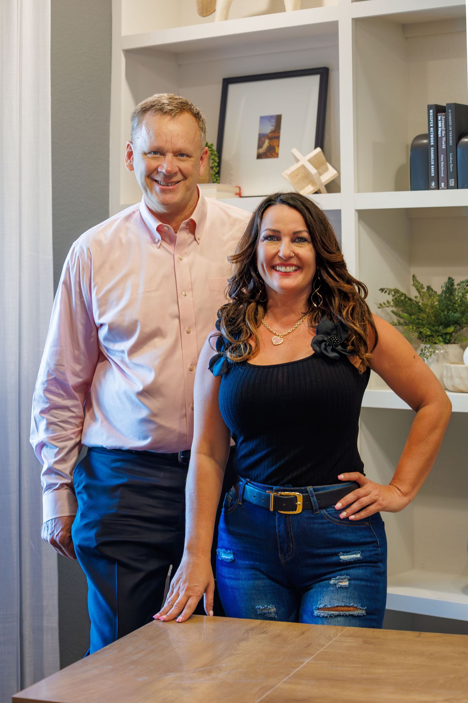 Two professionals stand together in an office with white built-in shelves, wearing business casual attire.
