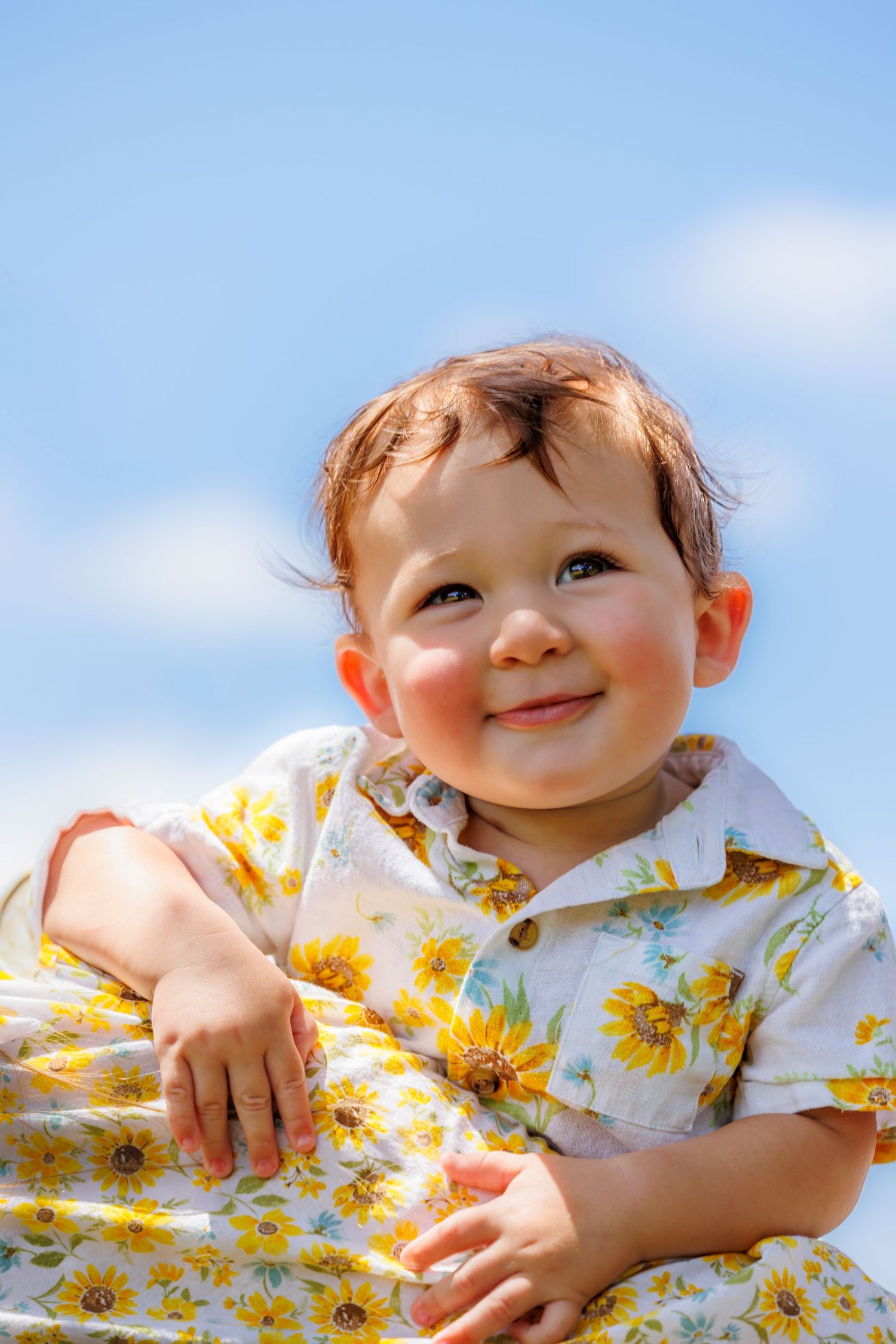 Young child in yellow floral outfit smiling against blue sky background.
