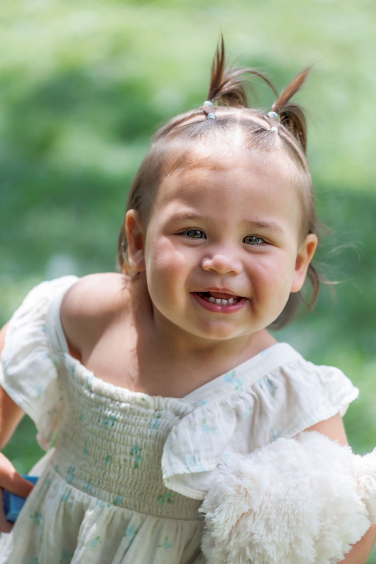 A smiling toddler wearing a white smocked dress with pigtails against a green background.