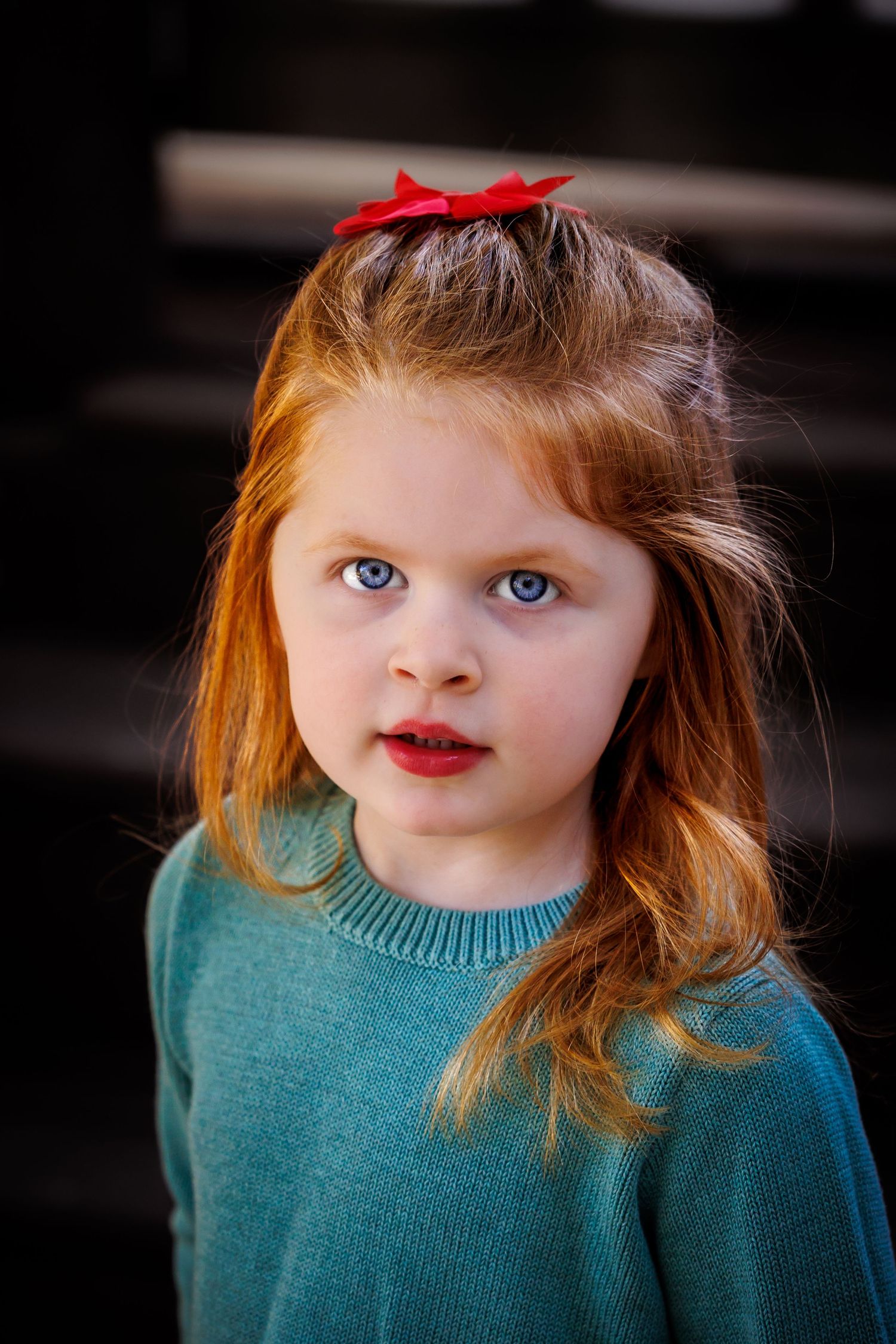 Portrait of a young child wearing a teal sweater and red bow against a dark background.