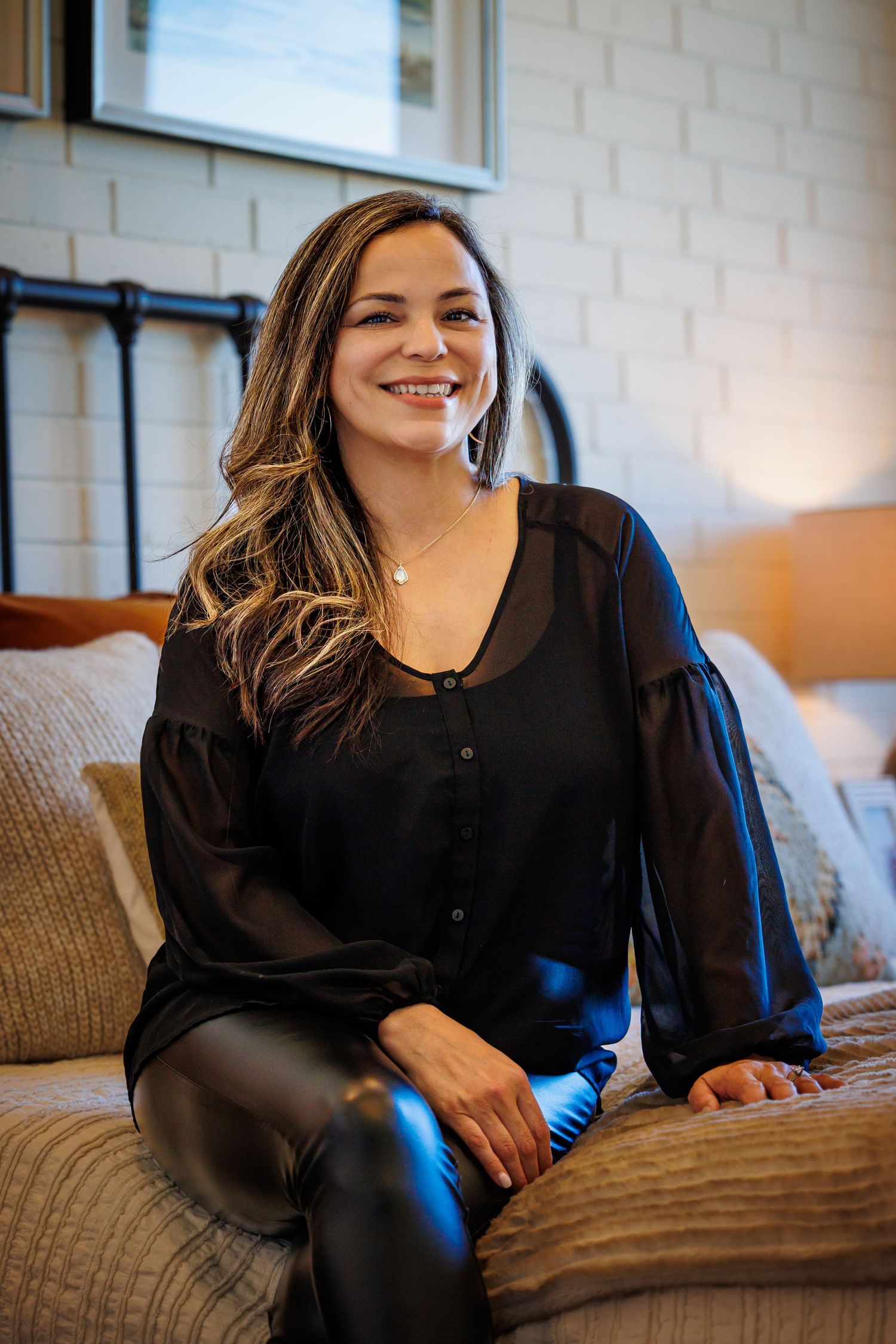 A person in a black sweater sits comfortably on a couch while smiling and looking to the side.
