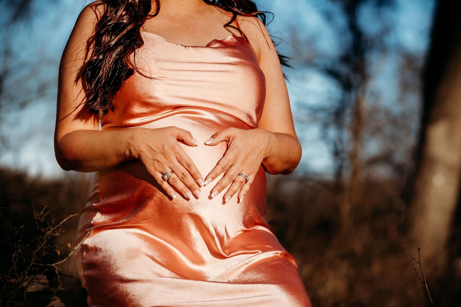 A person wearing a peach-colored silk dress poses outdoors with hands on their stomach in warm sunlight.