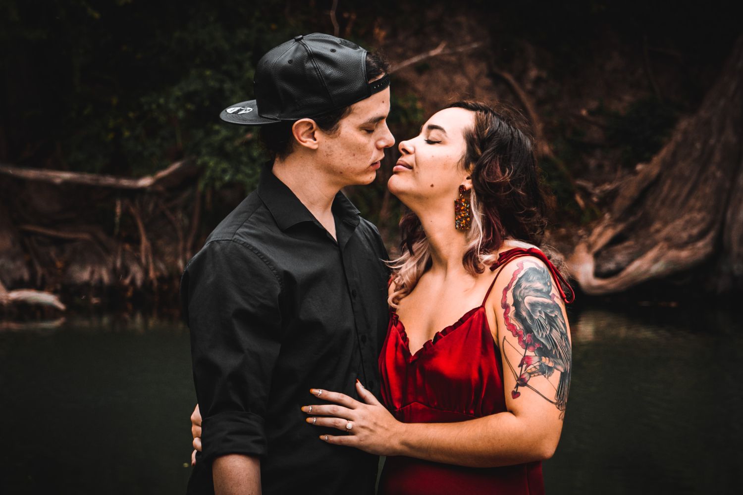 Romantic couple in red dress and black shirt embrace near water.