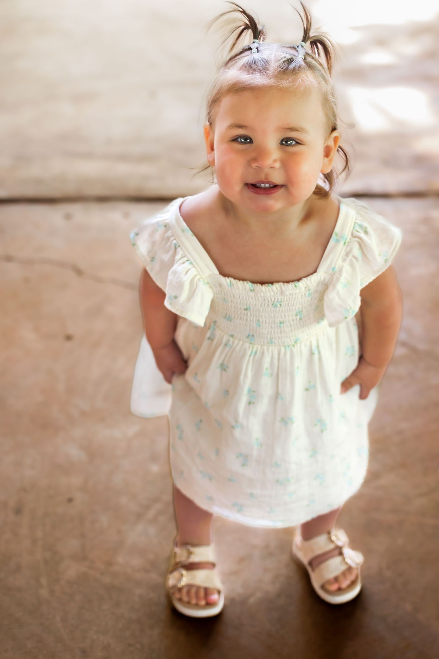 Toddler in white ruffled dress and sandals smiling at camera while standing indoors.