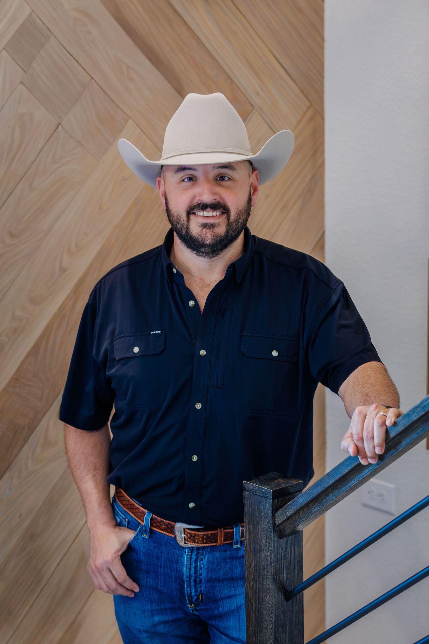 A person wearing a white cowboy hat, black button-down shirt and jeans stands near a wooden wall and staircase.